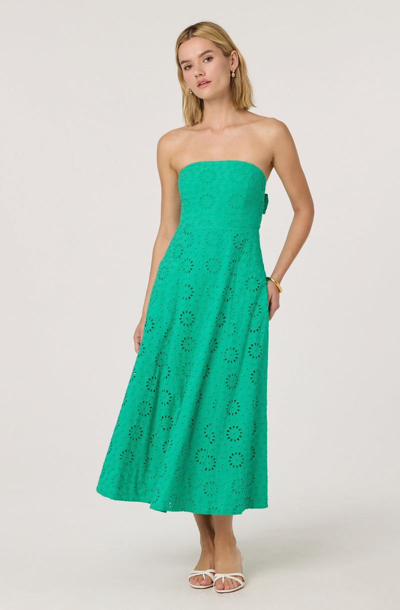A woman is wearing a strapless, turquoise dress with a eyelet pattern featuring circular and floral cutouts. The dress is fitted at the top and flows down to a midi length. She is accessorizing with earrings and a bracelet.