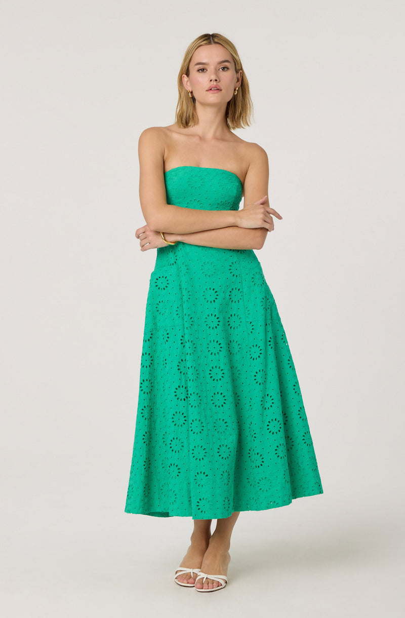 A woman is wearing a strapless, knee-length dress made of bright turquoise eyelet fabric with a circular floral pattern. The dress has a fitted bodice and a full, flowing skirt. She pairs it with white open-toe heels.