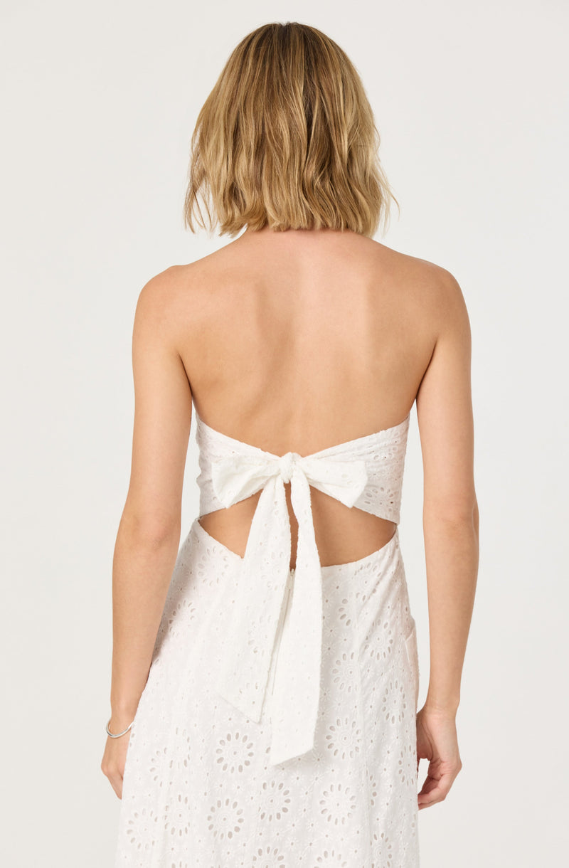 A woman is wearing a white, strapless dress with eyelet embroidery. The dress features a large bow tied at the back, creating cut-out sections on each side of the waist. The fabric has a floral eyelet pattern, giving it a textured appearance.