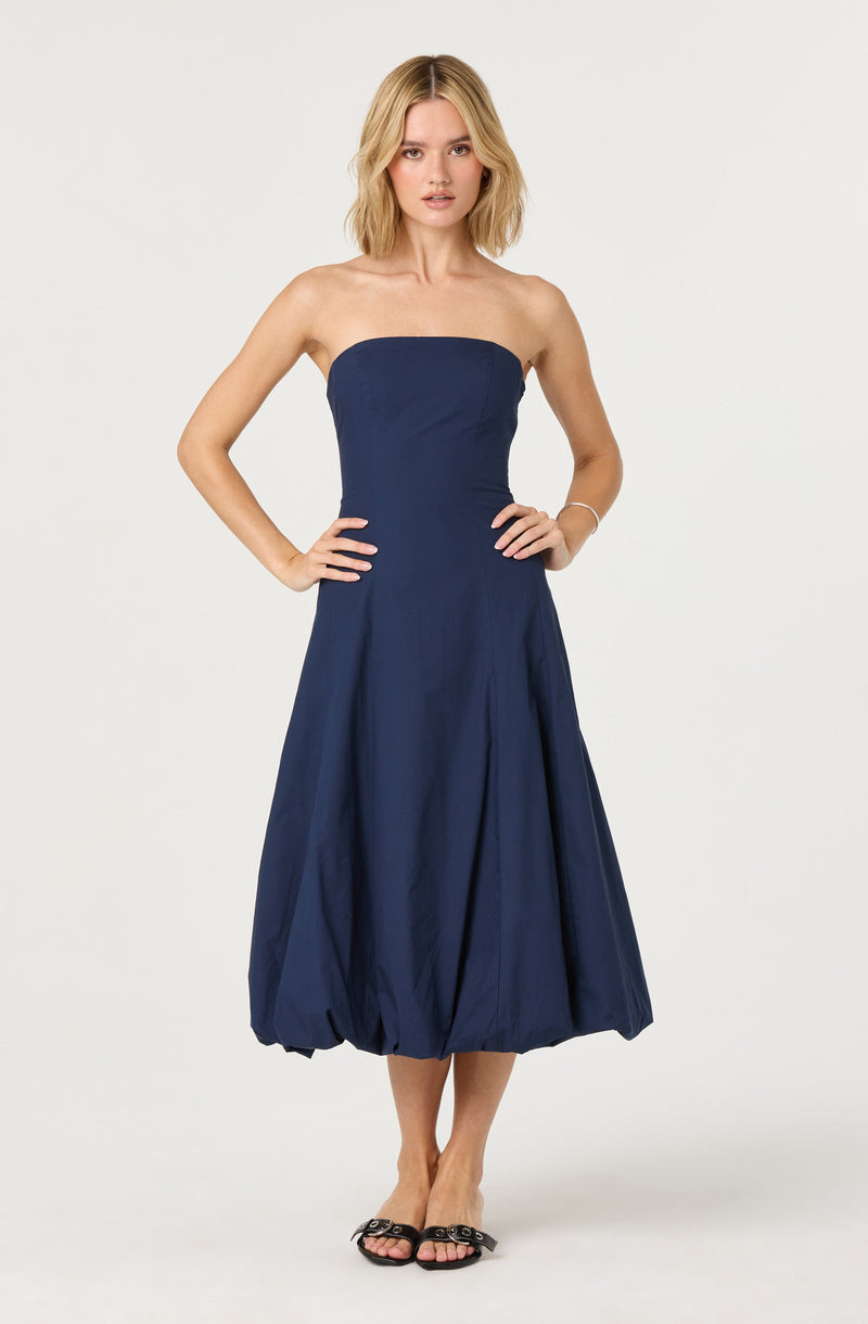 A woman is wearing a strapless, navy blue dress with a fitted bodice and a voluminous, bubble-style skirt that falls below the knees. She is also wearing black open-toe sandals with decorative embellishments.