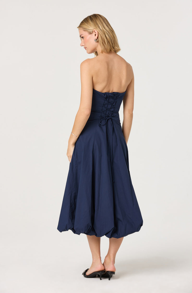 A woman is wearing a strapless, navy blue dress with a fitted bodice featuring lace-up detailing at the back. The dress has a full, balloon-style skirt that reaches mid-calf and is gathered at the hem. She is also wearing black high-heeled shoes and subtle jewelry, including earrings and a bracelet.