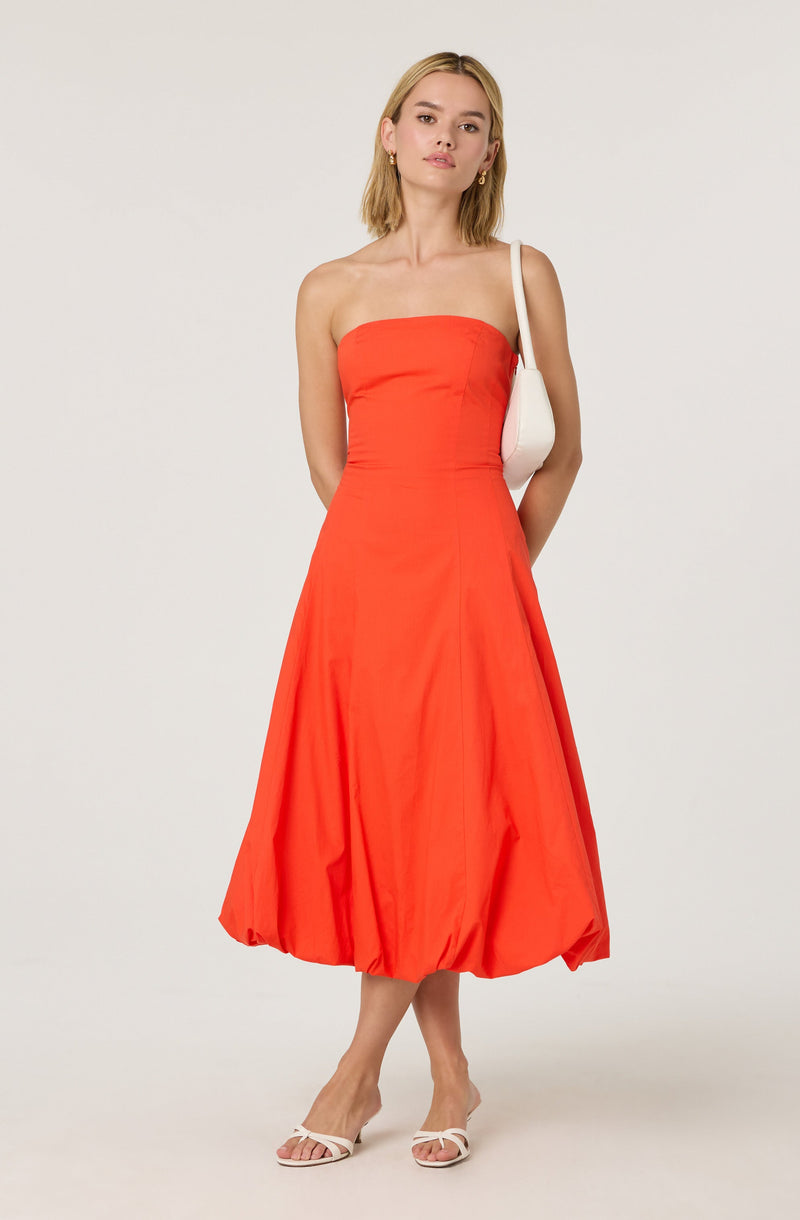 A woman is wearing a strapless, bright orange dress that has a fitted bodice and a gathered, voluminous skirt that reaches mid-calf length. She is carrying a white shoulder bag, wearing gold hoop earrings, and white open-toed heels.