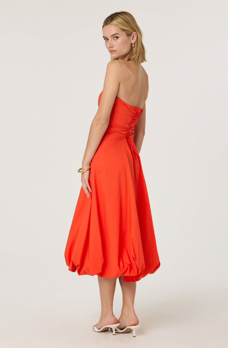 A woman is wearing a strapless, bright orange, midi-length dress with a fitted bodice featuring lace-up detail on the side. The skirt is voluminous with gathered, puffed sections at the hem. She has on white high-heeled shoes with open toes and ankle straps, and she is accessorizing with a gold bracelet and earrings.