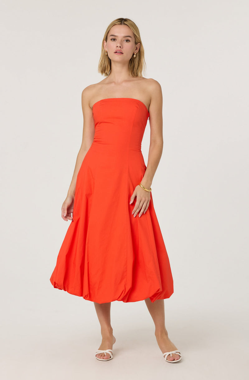 A woman is wearing a strapless, bright orange maxi dress with a fitted bodice and a voluminous, gathered skirt that stops mid-calf. She is accessorizing with a gold bracelet and small gold earrings. She has blonde hair styled in loose waves and is standing against a plain, light-colored background, wearing white open-toe heels.