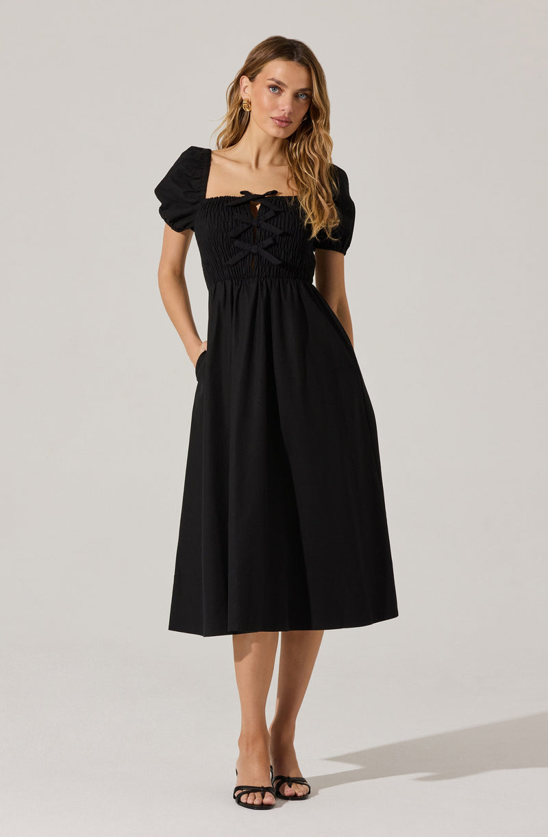 A woman is wearing a black midi dress with puffed short sleeves. The bodice features a smocked design with crisscross lace-up detailing at the neckline. The skirt is plain and flows loosely, reaching mid-calf. She is accessorizing with gold hoop earrings and black strappy high-heeled sandals.