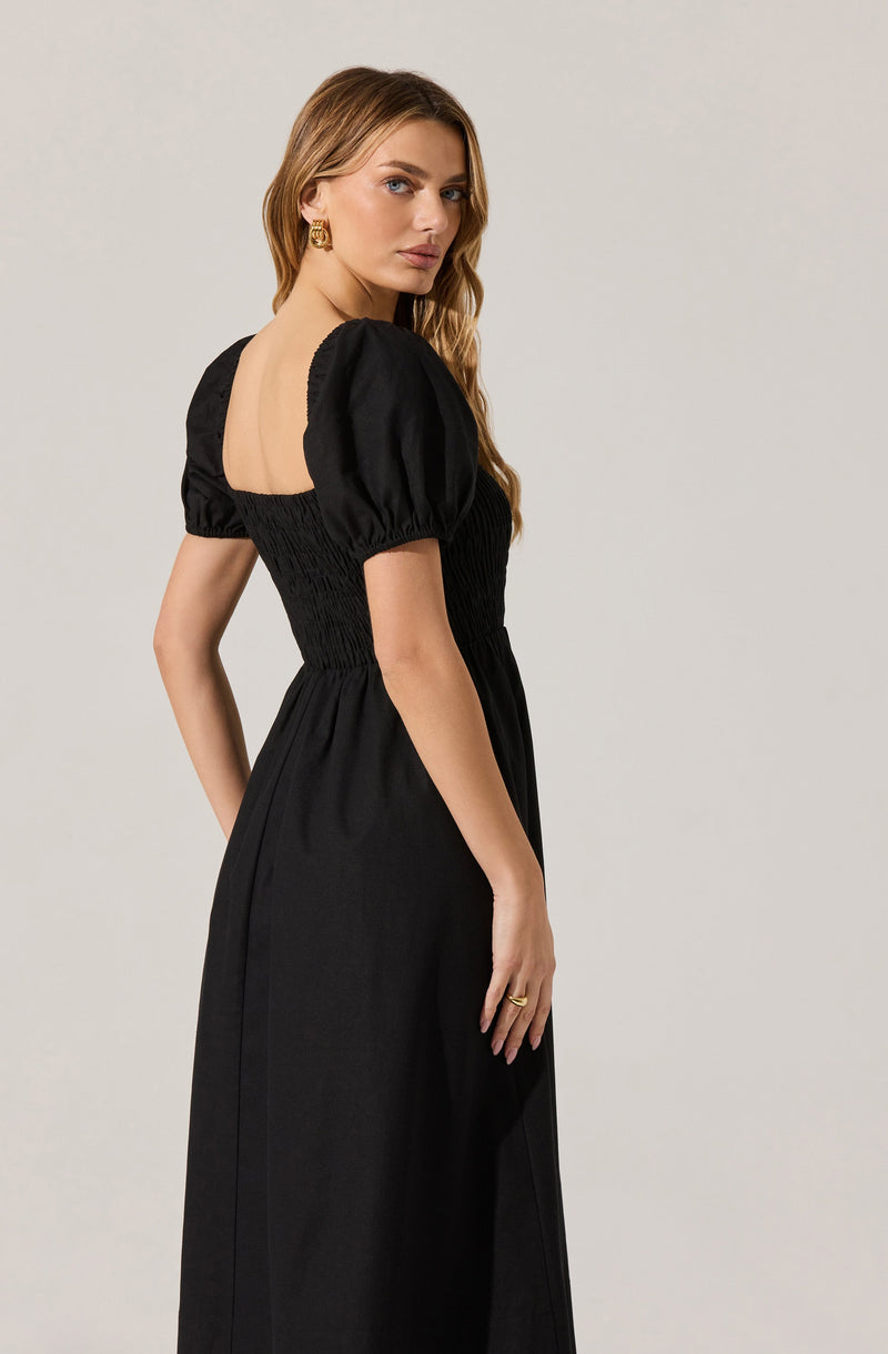 A woman is wearing a black dress with puffed short sleeves gathered at the shoulders and elasticated at the waist, creating a fitted bodice that flows into a long, straight skirt.