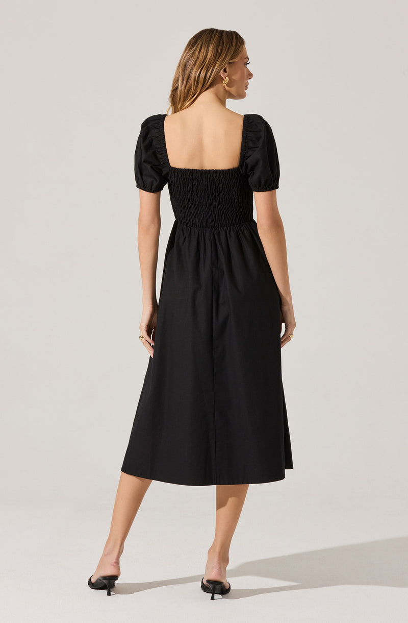A woman is wearing a black dress with a fitted, elasticized waist and a square neckline. The dress features short, puffed sleeves with ruching detail. She is also wearing black open-toe heels.