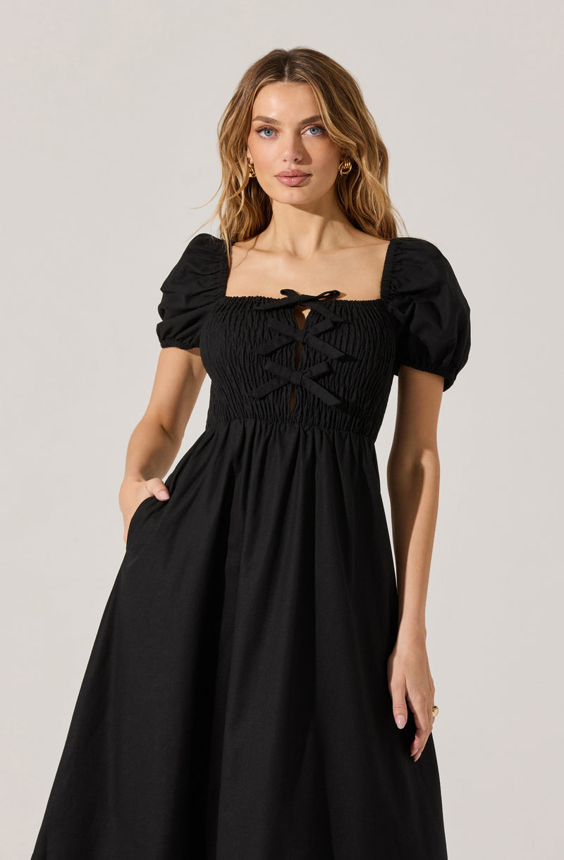 A woman is wearing a black maxi dress with puffed short sleeves. The bodice features ruched fabric with a lace-up detail in the front, creating a gathered, textured appearance. The skirt is long, flowing, and smooth, with the woman resting her hand in a pocket on the side.