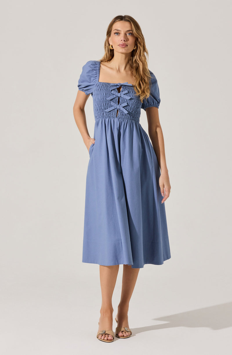A woman is wearing a light blue midi dress with puffed short sleeves and a square neckline. The bodice features smocking and decorative lacing with crisscrossed ties at the front. The skirt is gathered and flows loosely down to mid-calf length. She is also wearing beige heeled sandals.