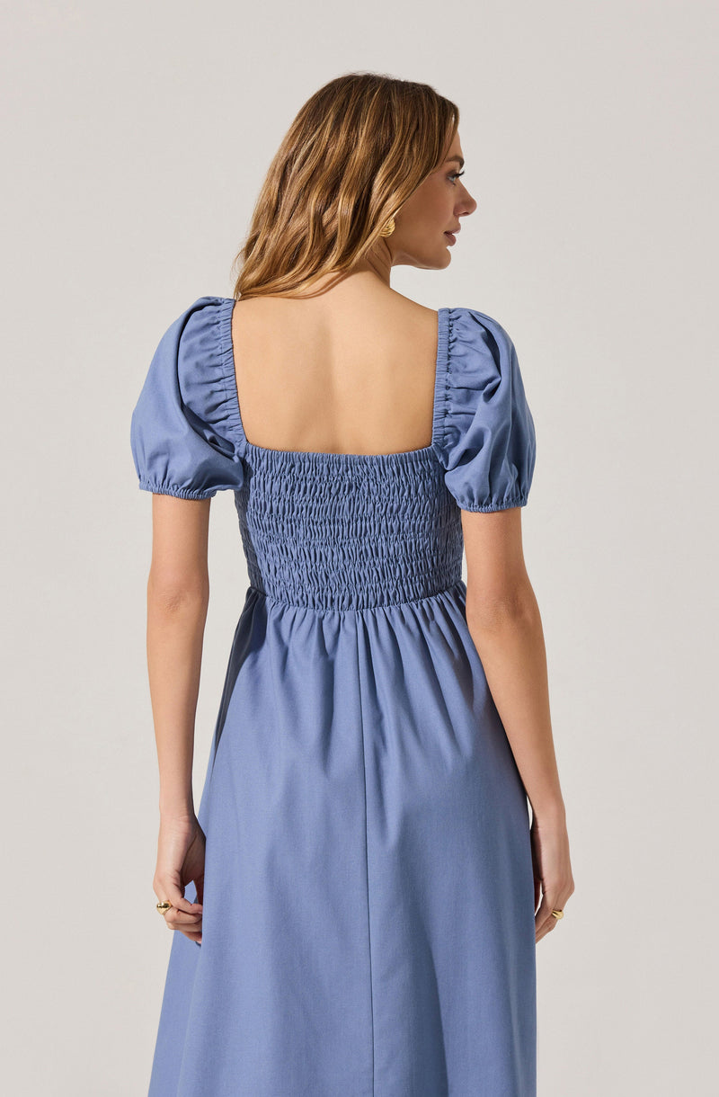 A woman is wearing a light blue dress with short, puffed sleeves that have gathered elastic at the edges. The dress features a wide, square neckline in the back, with a smocked, fitted bodice that creates a textured, ruched appearance. The skirt of the dress is flowy and falls loosely.
