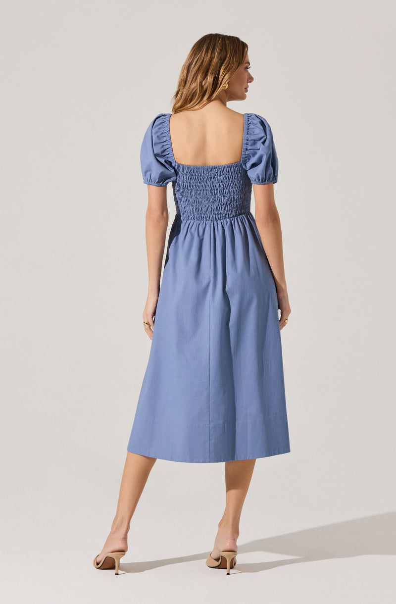 A woman is wearing a light blue midi dress with puffed short sleeves and a square neckline. The dress features a smocked bodice that extends to the waist, creating a fitted appearance, and the skirt is gathered, flowing loosely to mid-calf length. She is also wearing nude high-heeled shoes.