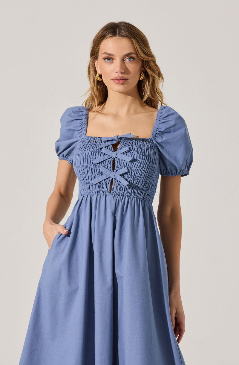 A woman is wearing a light blue dress with short puffed sleeves. The dress features a square neckline with a smocked bodice that has a lace-up detail at the front, including multiple matching blue ribbon ties. The skirt is gathered at the waist and flows down in a loose, flowing style.