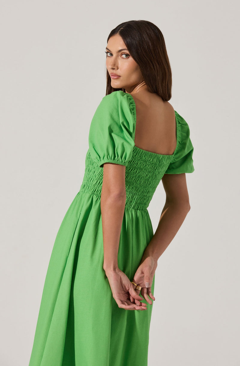 A woman is wearing a bright green dress with puffed short sleeves. The bodice of the dress features shirred or ruched detailing, creating a fitted waist, and the skirt is flowing and long. The dress has a wide, square neckline that exposes her shoulders and upper back.