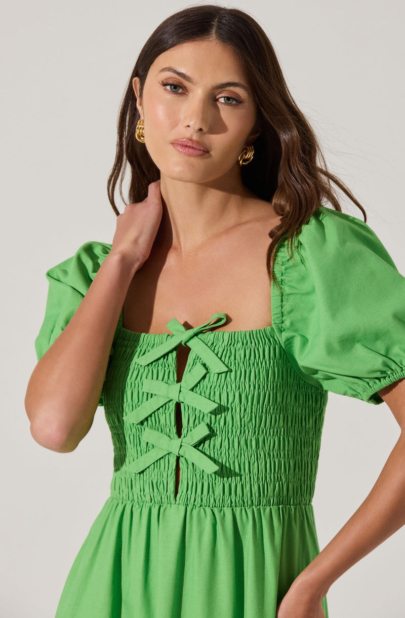 A woman is wearing a bright green dress with short puff sleeves. The bodice features ruched fabric with a lace-up detail in the center, tied into a bow at the neckline. The skirt portion of the dress is gathered and flows loosely. She is also wearing gold hoop earrings.