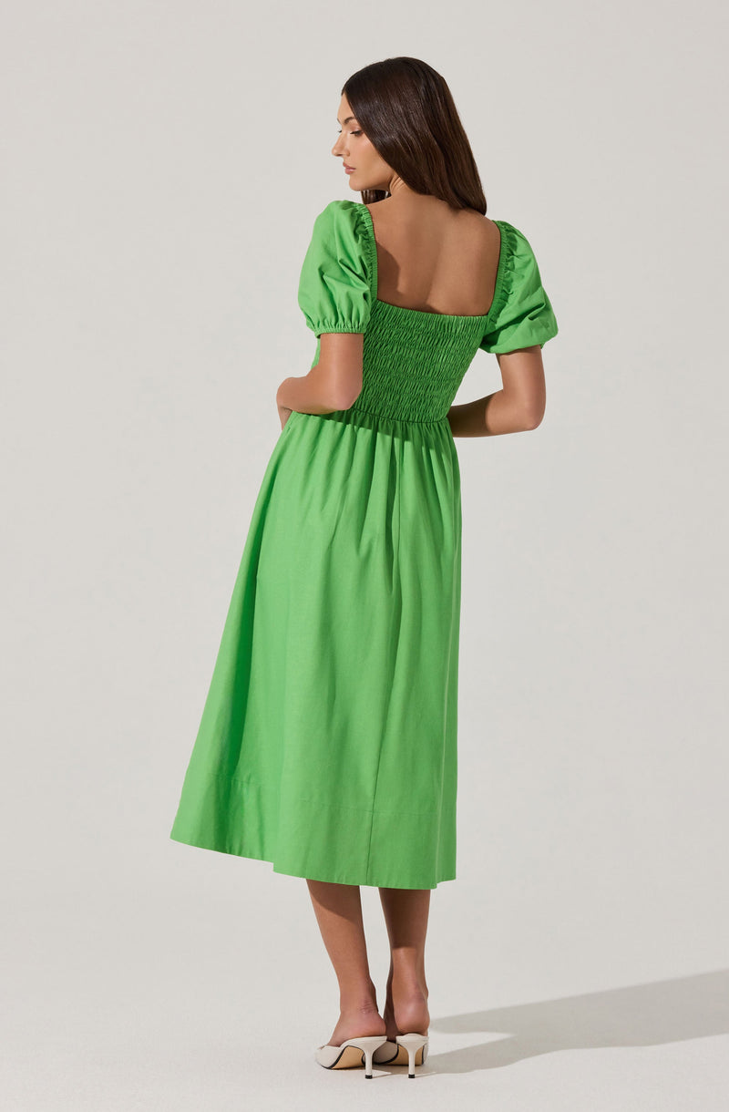 A woman is wearing a bright green, midi-length dress with puffed short sleeves and a smocked bodice. The dress has a square neckline at the back and flows loosely down to her ankles. She is also wearing light-colored high-heeled shoes.