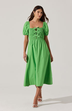 A woman is wearing a bright green midi dress with a square neckline and short, puffed sleeves. The bodice features smocking and decorative crisscross laces at the front. She pairs the dress with open-toe heels and gold earrings.