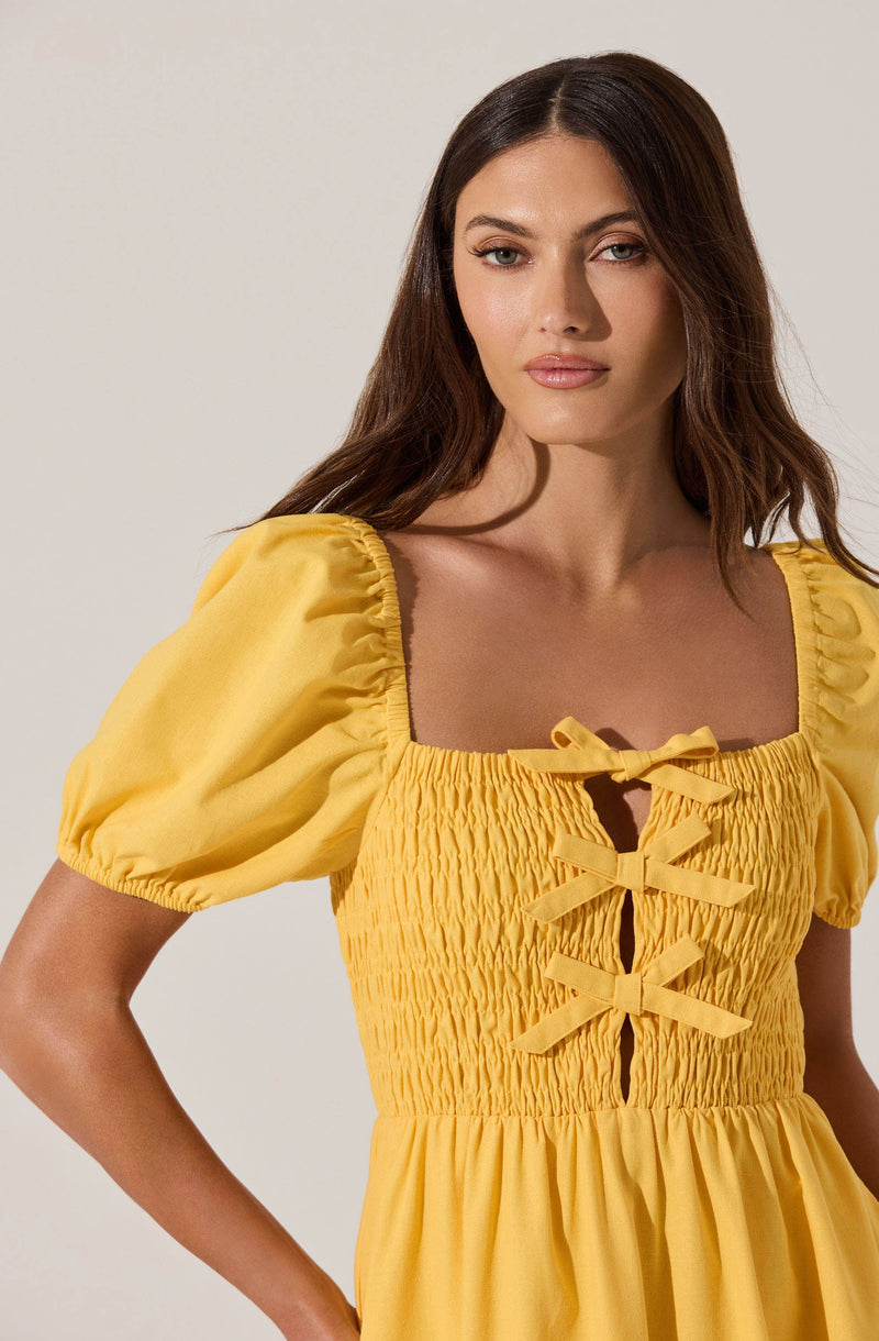 A woman is wearing a bright yellow dress with puffed short sleeves. The bodice features a smocked, ruched design with a square neckline. There are three decorative bows in matching yellow fabric arranged vertically along the front center of the dress. The skirt portion of the dress is gathered and flows loosely.