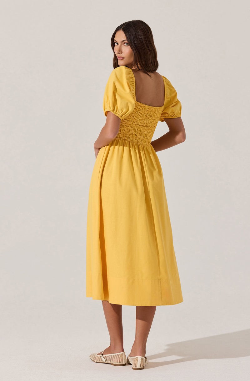 A woman is wearing a yellow dress with puffed short sleeves that have elasticized cuffs. The dress features a fitted, ruched bodice at the back with a square neckline. The skirt of the dress is long and flowing, reaching mid-calf. She is also wearing beige open-toe shoes with a woven texture.