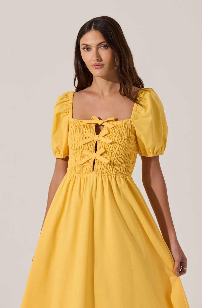 A woman is wearing a bright yellow dress with puffed short sleeves and a square neckline. The bodice features smocking and decorative bows running down the front. The skirt is full and gathered, creating a flowing silhouette.