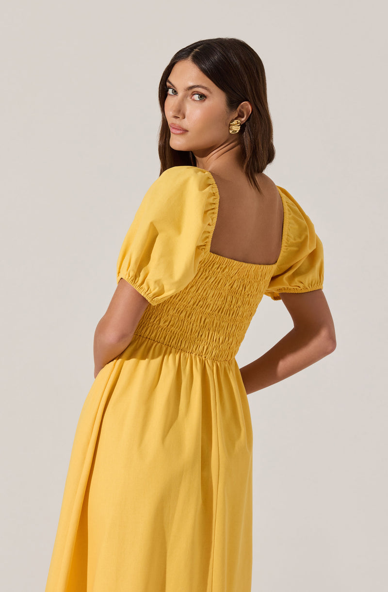 A woman is wearing a bright yellow dress with puffed short sleeves. The dress has a square neckline in the back with smocked detail around the waist, creating a fitted look. The skirt of the dress is full and gathered, flowing down to the floor. She is accessorized with gold earrings.