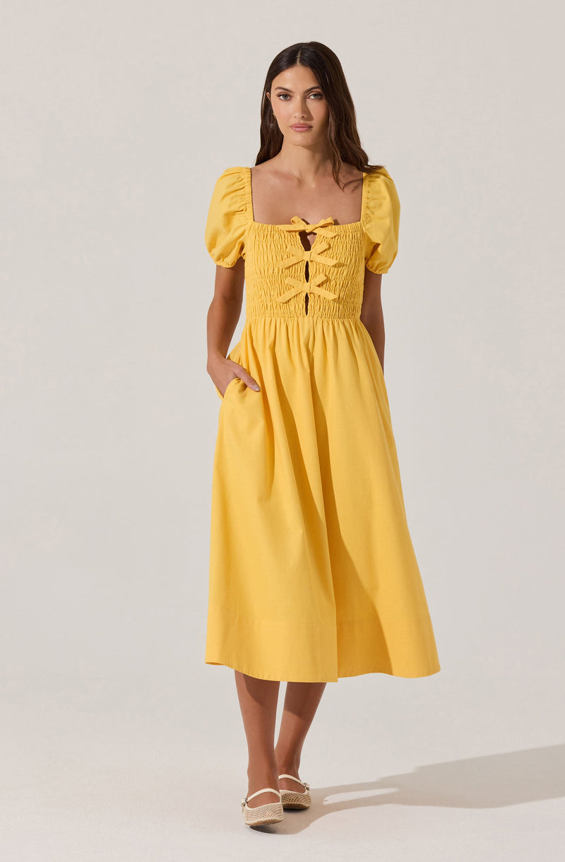 A woman is wearing a bright yellow dress with puffed short sleeves and a square neckline. The bodice features smocking and decorative ribbon lacing at the front. The dress has a fitted waist and a full, flowing skirt that reaches mid-calf length. She is wearing beige shoes with ankle straps.