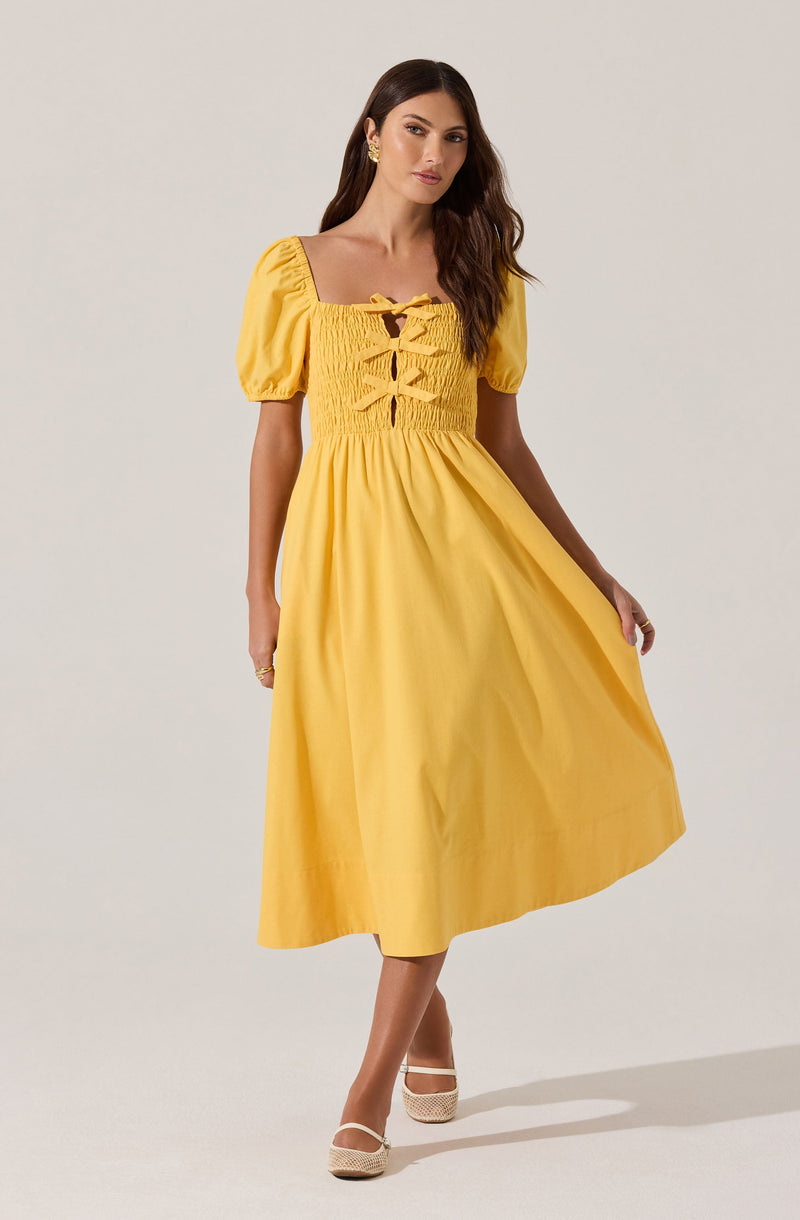 The woman is wearing a yellow, ankle-length dress with a fitted, smocked bodice featuring a lace-up detail at the front. The dress has short, puffed sleeves and a full, flowing skirt. She is also wearing white, closed-toe shoes with ankle straps.