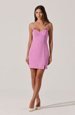 A woman is wearing a fitted, sleeveless pink dress with thin spaghetti straps. The straps feature small knot bows near the shoulders. The dress has a structured bodice with visible seam details and a slight slit on the side hem. She is also wearing white high-heeled shoes.