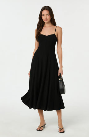A woman is wearing a black, sleeveless midi dress with thin spaghetti straps and a fitted bodice. The dress has a flared skirt that falls below the knees. She is also holding a small black handbag with a gold accent, and wearing black open-toe high-heeled sandals.