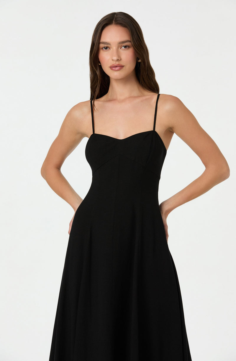 A woman is wearing a long, black dress with spaghetti straps, which features a fitted bodice and a flowing skirt. The dress has a sweetheart neckline and appears to be made from a smooth, elegant fabric.
