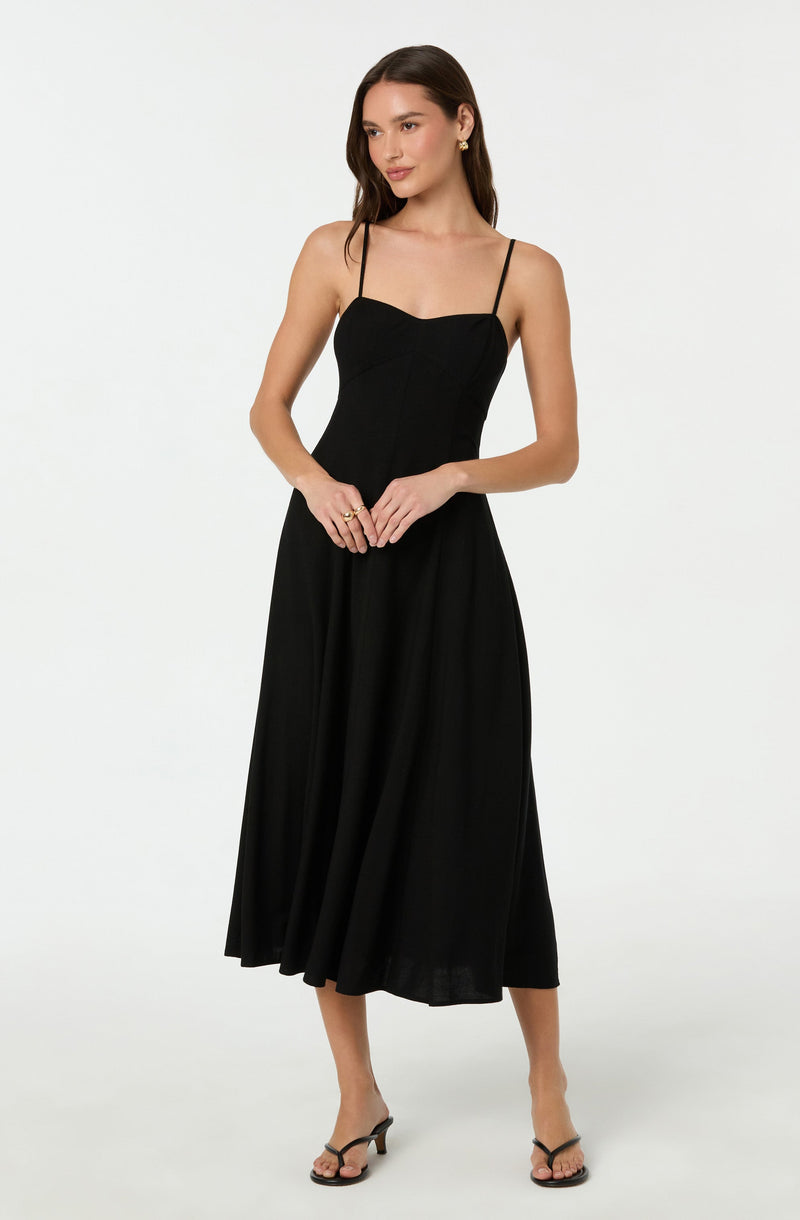 A woman is wearing a black sleeveless dress with thin spaghetti straps, a fitted bodice, and a flowing, ankle-length skirt. She is also wearing black high-heeled sandals.