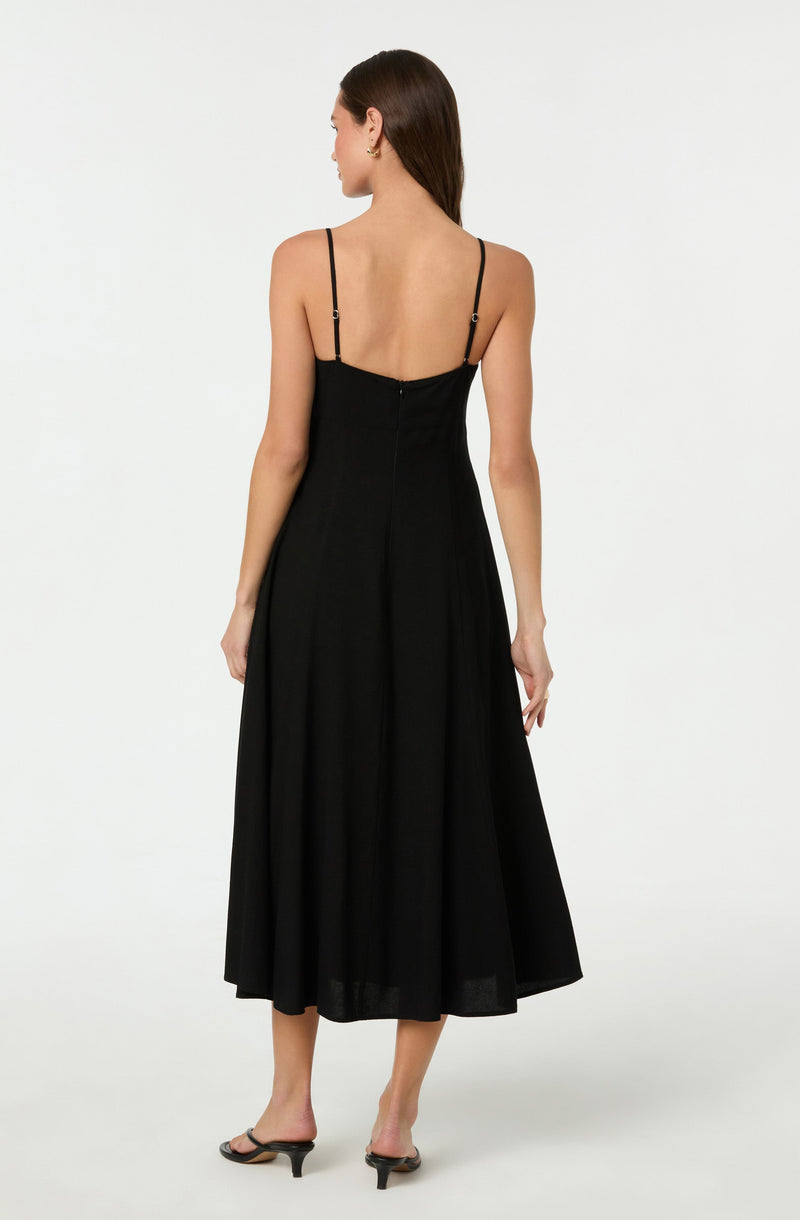 A woman is wearing a black spaghetti strap dress that falls to mid-calf length. The dress has a fitted bodice with thin adjustable straps and a slightly flared skirt. She is also wearing black open-toed heels with a small heel.