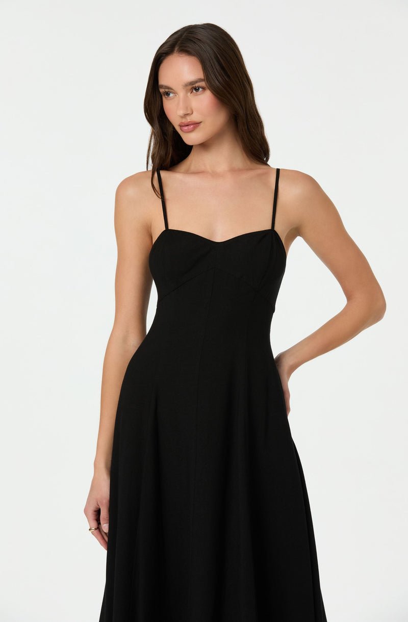 A woman is wearing a black sleeveless dress with thin, adjustable spaghetti straps. The dress features a fitted bodice with subtle seaming details and a flowing, floor-length skirt.