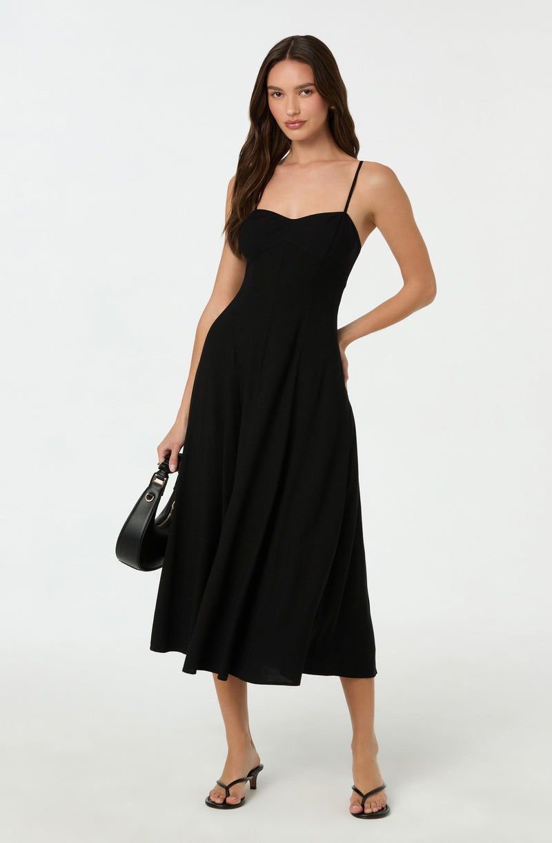 A woman is wearing a black midi dress with thin spaghetti straps and a fitted bodice. She is also holding a small black handbag and wearing black strappy high-heeled sandals.
