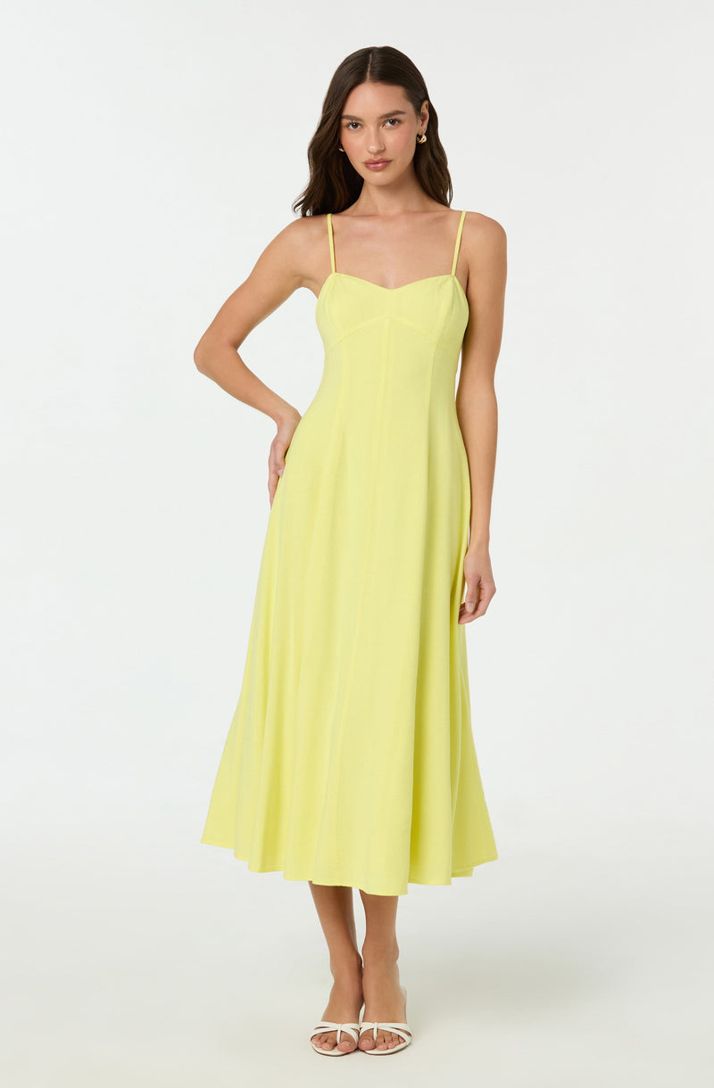 A woman is wearing a sleeveless, ankle-length dress with thin spaghetti straps. The dress is a solid, light yellow color and features a fitted bodice with a sweetheart neckline. The skirt flows loosely to the ankles, creating an elegant silhouette. She is also wearing white heeled sandals.