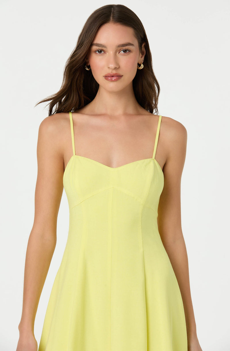 A woman is wearing a sleeveless, yellow dress with thin spaghetti straps. The dress features a fitted bodice with a sweetheart neckline and a slightly flared skirt.