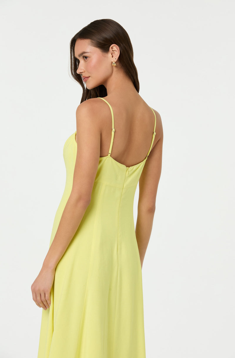 A woman is wearing a sleeveless, yellow dress with thin shoulder straps. The back of the dress features a zipper closure, and the fabric appears lightweight and flowy. She has long, dark hair and is accessorized with small, gold earrings.
