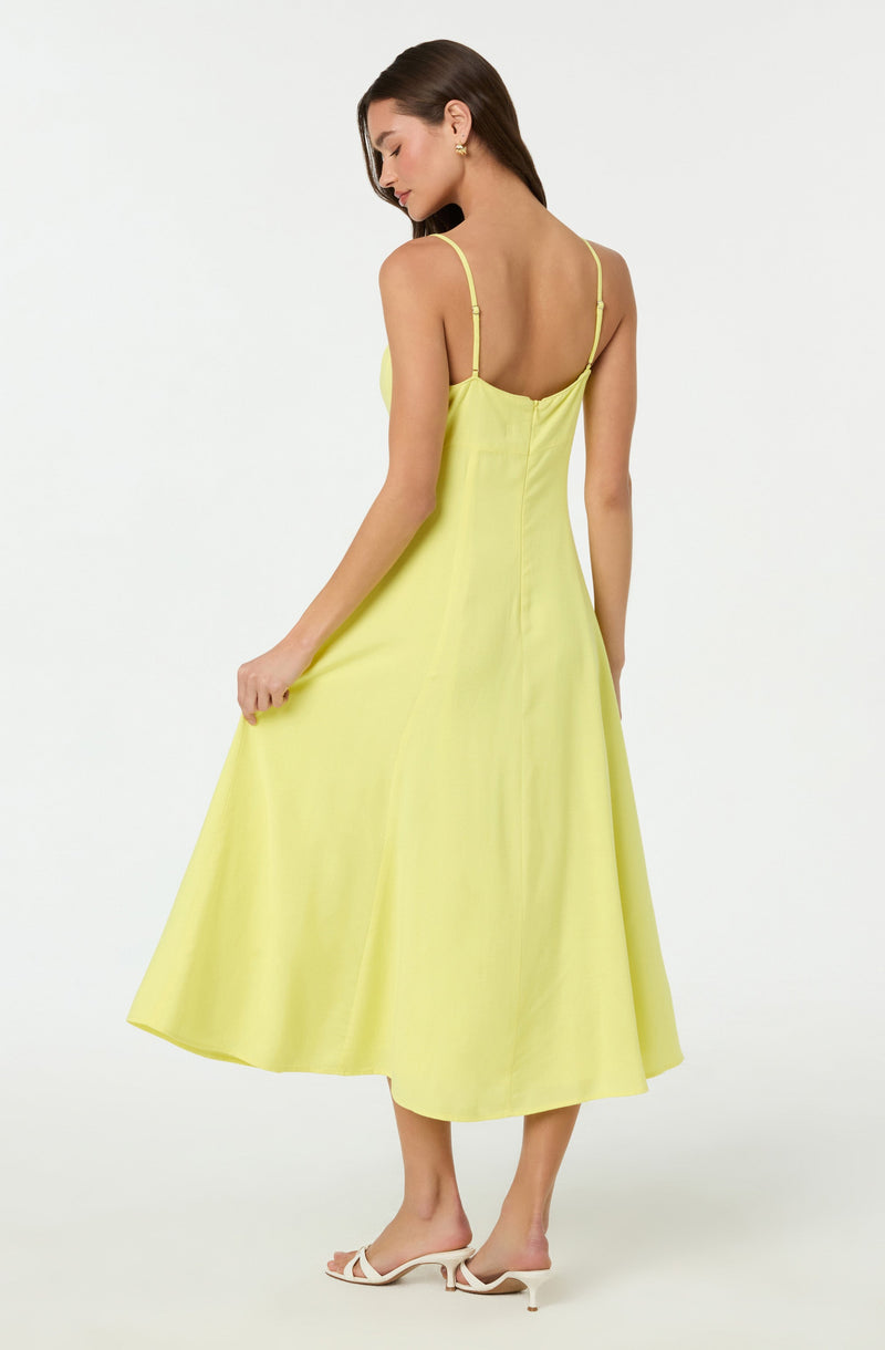 A woman is wearing a bright yellow midi dress with thin adjustable spaghetti straps. The dress has a fitted bodice and a flared skirt that reaches mid-calf. She pairs the dress with white open-toe high-heeled sandals.