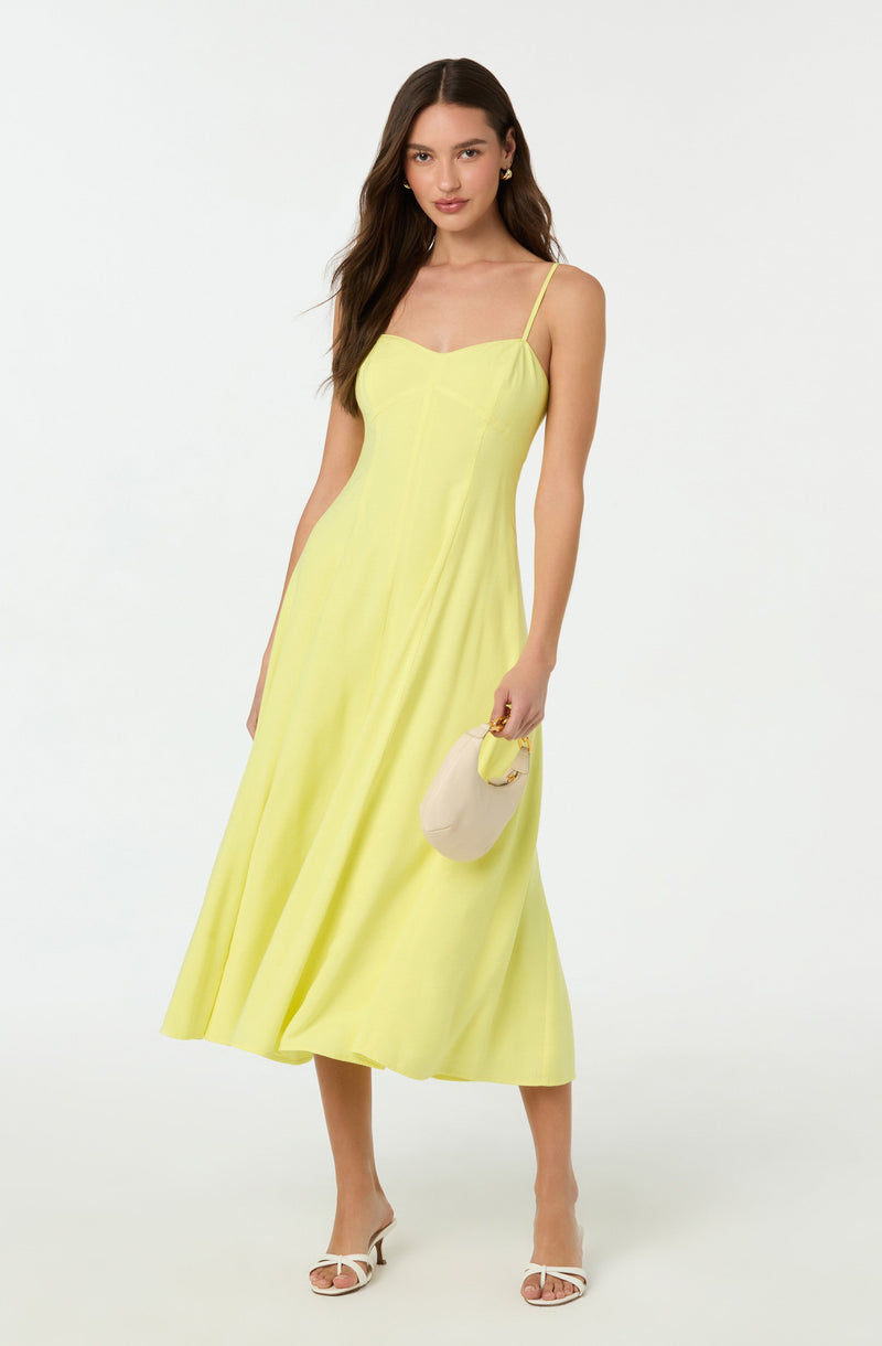 A woman is wearing a yellow sleeveless dress with thin spaghetti straps and a fitted bodice. The dress is long, flowing, and reaches down to her ankles. She is holding a light-colored, rounded handbag with a yellow handle in her right hand. She is also wearing white open-toe high heels with a slim strap across the toes.