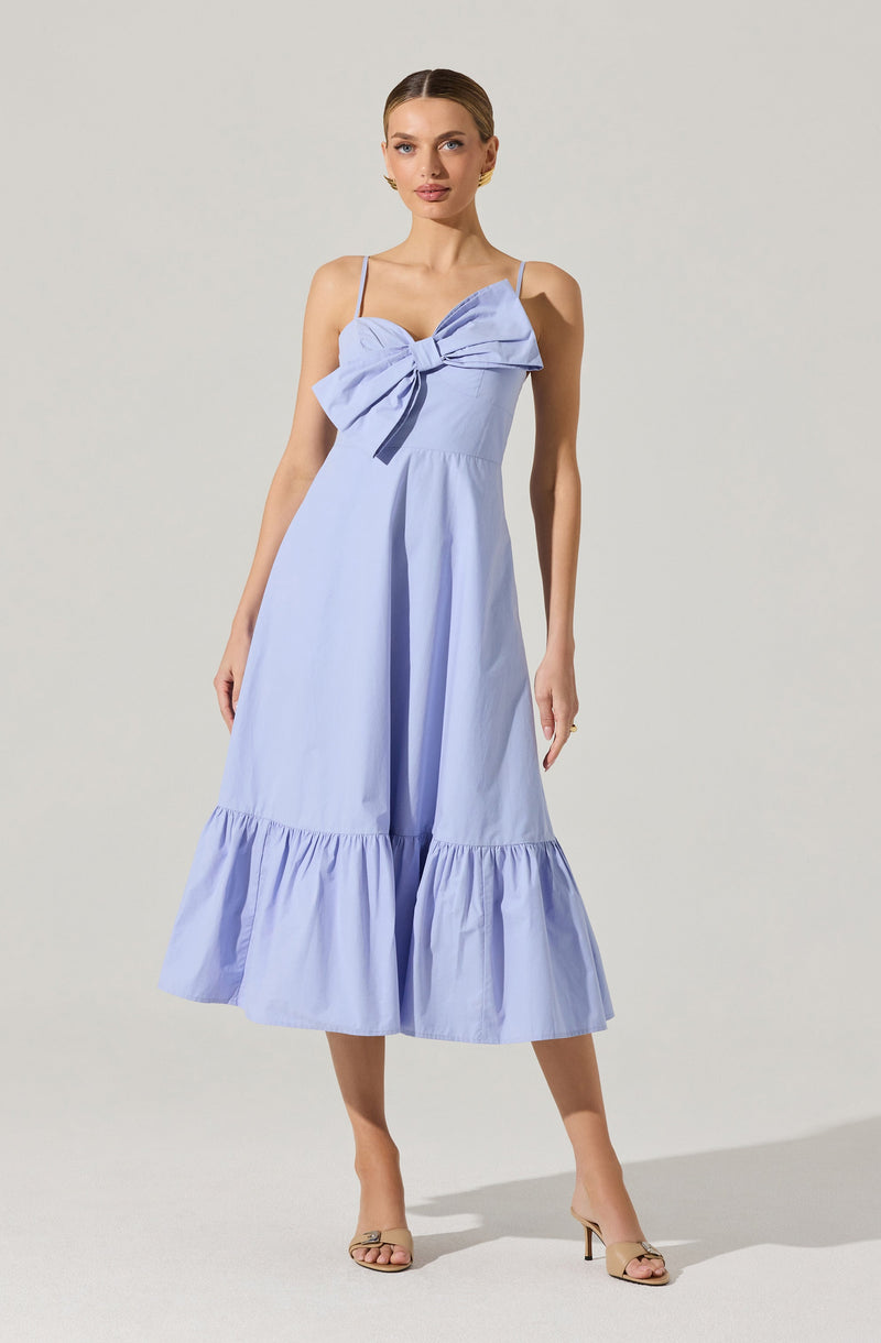 A woman is wearing a light blue spaghetti strap dress featuring a large decorative bow on the chest area. The dress has a fitted bodice and a full, A-line skirt that reaches mid-calf, with a ruffled hem at the bottom. The fabric appears to be lightweight and slightly structured. She is also wearing open-toe heels with a thin strap around the ankle.