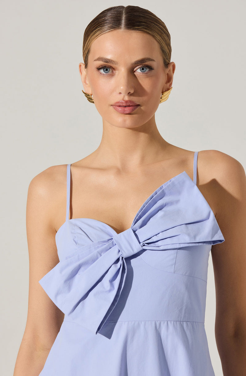A woman is wearing a light blue dress with thin spaghetti straps. The dress features a large, prominent bow on the front, made from the same fabric as the dress.