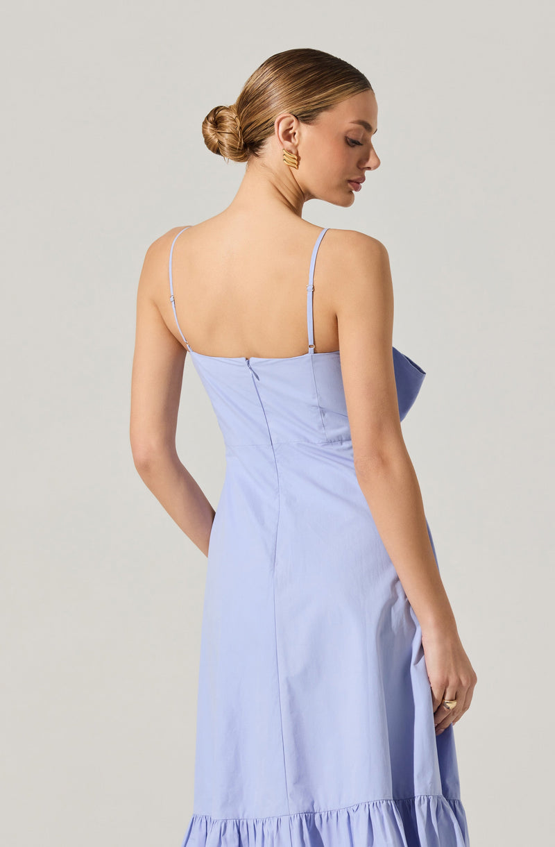 A woman is wearing a light blue sundress with thin spaghetti straps. The dress features a fitted bodice and a ruffled hem at the bottom. She is accessorized with gold hoop earrings and has her hair styled in a low, twisted bun.