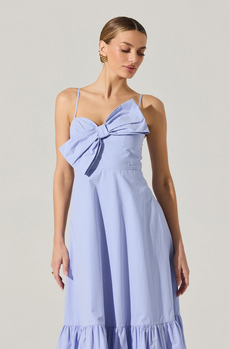 A woman is wearing a light blue, sleeveless dress with thin spaghetti straps. The dress features a large, decorative bow on the front left side of the bodice, made from matching fabric. The skirt of the dress is long with a gathered, ruffled hemline at the bottom.