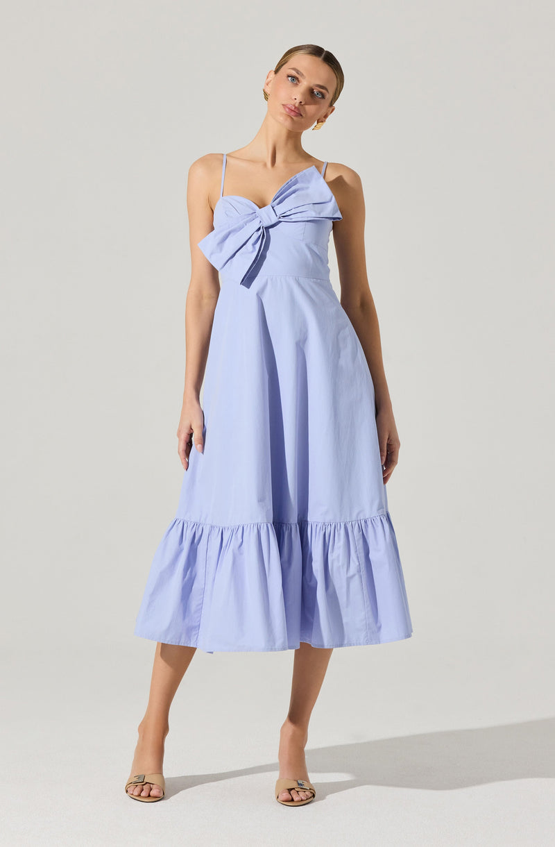 A woman is wearing a light blue, sleeveless, midi-length dress with thin spaghetti straps. The dress features a large bow with a knot at the chest area, creating a draped effect. The skirt has a gathered ruffle hem at the bottom, adding volume and a playful touch to the overall design.