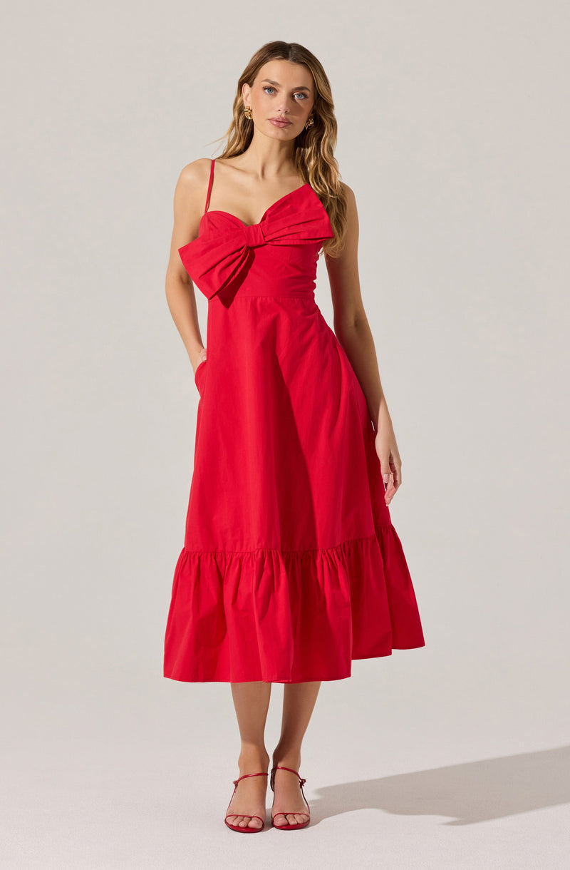A woman is wearing a sleeveless, red midi-length dress with spaghetti straps. The dress features a large bow on the bust area, creating a decorative detail. The skirt has a ruffled hemline that adds volume and movement to the dress. She completes her outfit with matching red strappy high-heeled sandals.