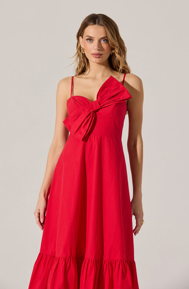 A woman is wearing a sleeveless, red, ankle-length dress with thin spaghetti straps. The dress features a large, prominent bow on the left shoulder. The skirt has a gathered, ruffled hem at the bottom.