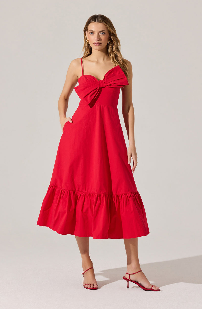 A woman is wearing a sleeveless red dress with thin spaghetti straps. The dress features a large bow detail at the bust area, and has a fitted waist. The skirt is midi length with a tiered ruffle at the hem. She is also wearing red high-heeled sandals with thin straps.