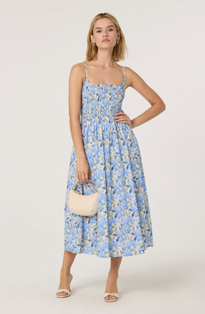 A woman is wearing a light blue sleeveless dress with thin spaghetti straps, featuring a floral pattern of blue and beige leaves. She is accessorizing with small gold earrings and holding a small, cream-colored handbag. She is also wearing beige open-toe heels.