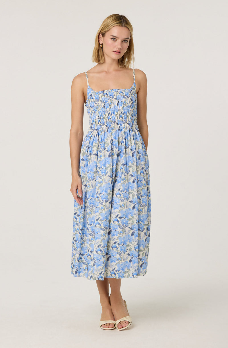 A woman is wearing a sleeveless, ankle-length dress with thin spaghetti straps. The dress features a fitted, smocked bodice and a flowing skirt with a floral pattern in shades of blue, beige, and light lavender. She is also wearing open-toed, light-colored high-heeled sandals.
