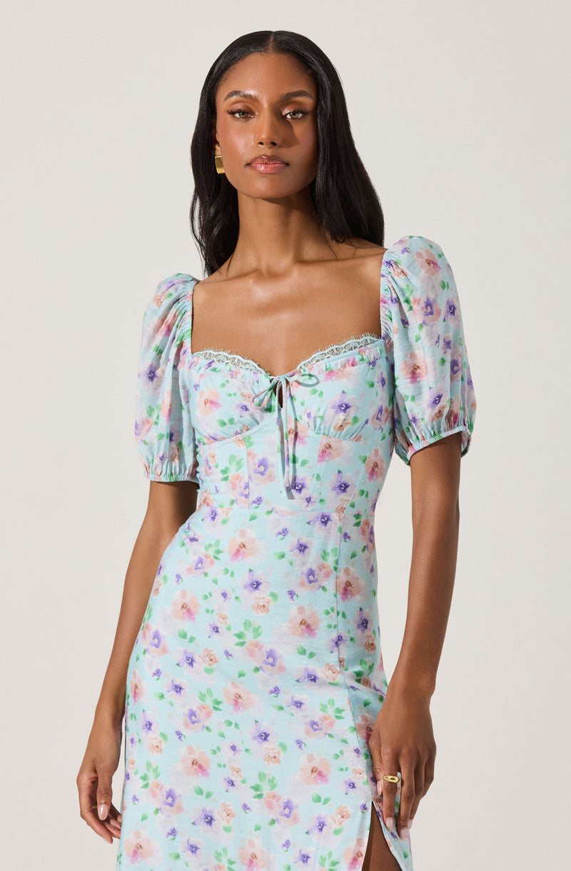A woman is wearing a light-colored dress with a pastel floral print featuring purple, pink, and green hues. The dress has puffed short sleeves, a square neckline with lace trim, and a tie detail at the bust.