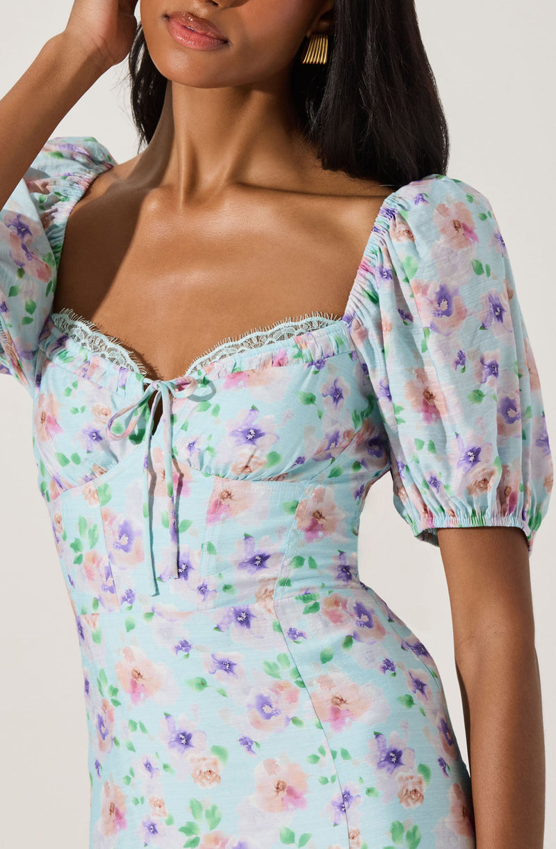 A woman is wearing a fitted dress with a pastel floral pattern featuring pink, purple, and green colors on a light background. The dress has puffed short sleeves gathered at the shoulders, a square neckline trimmed with white lace, and a small bow tie at the center of the neckline. The fabric appears lightweight and soft.